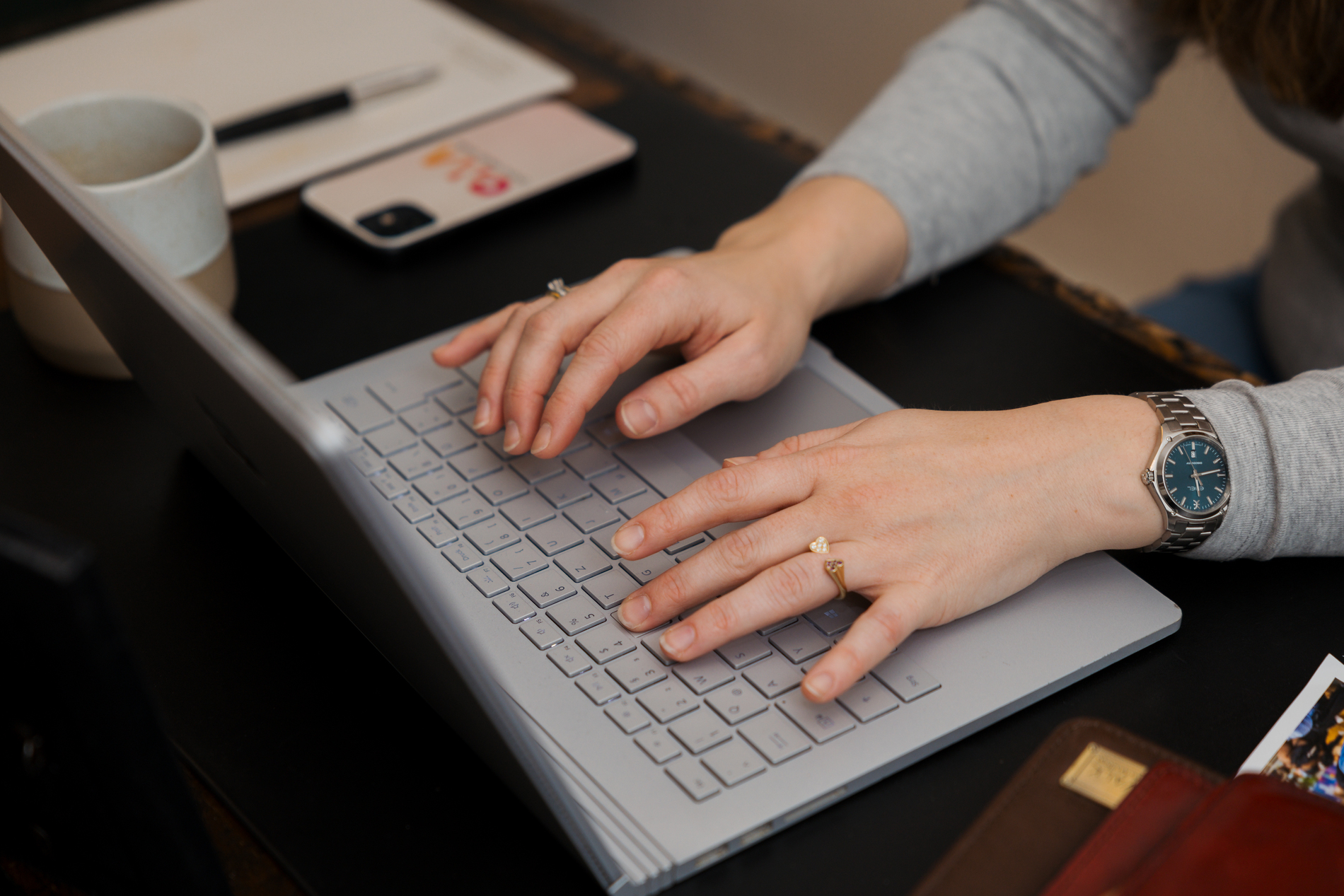 Person tippt auf Laptop-Tastatur, daneben liegen Smartphone, Tasse und Notizbuch auf einem Schreibtisch.